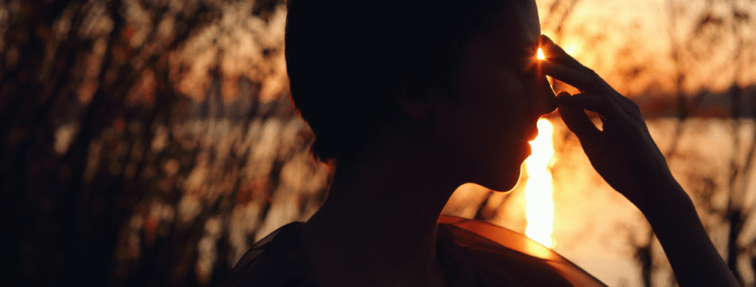 A woman holding her fingers to her third eye with the setting sun shining through her fingers